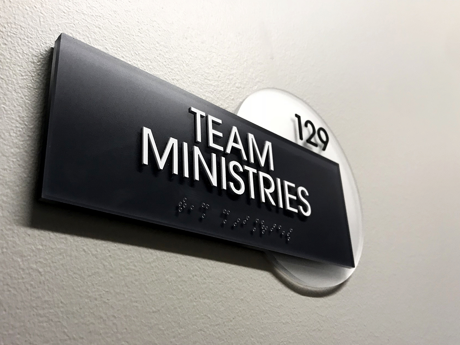 Illuminated sign with braille and text for Team Ministries, mounted on interior wall.