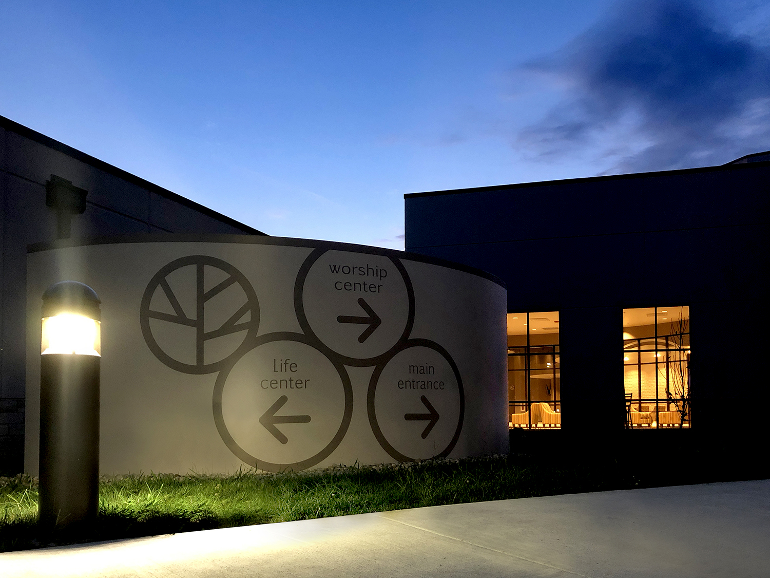 Modern church sign with directional arrows for worship, life, and main entrance at dusk.