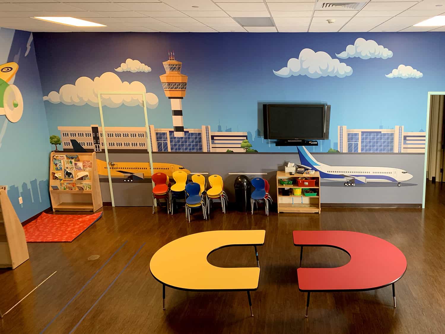 Bright children's playroom with colourful seating and a city-themed wall mural featuring an airport and airplane.