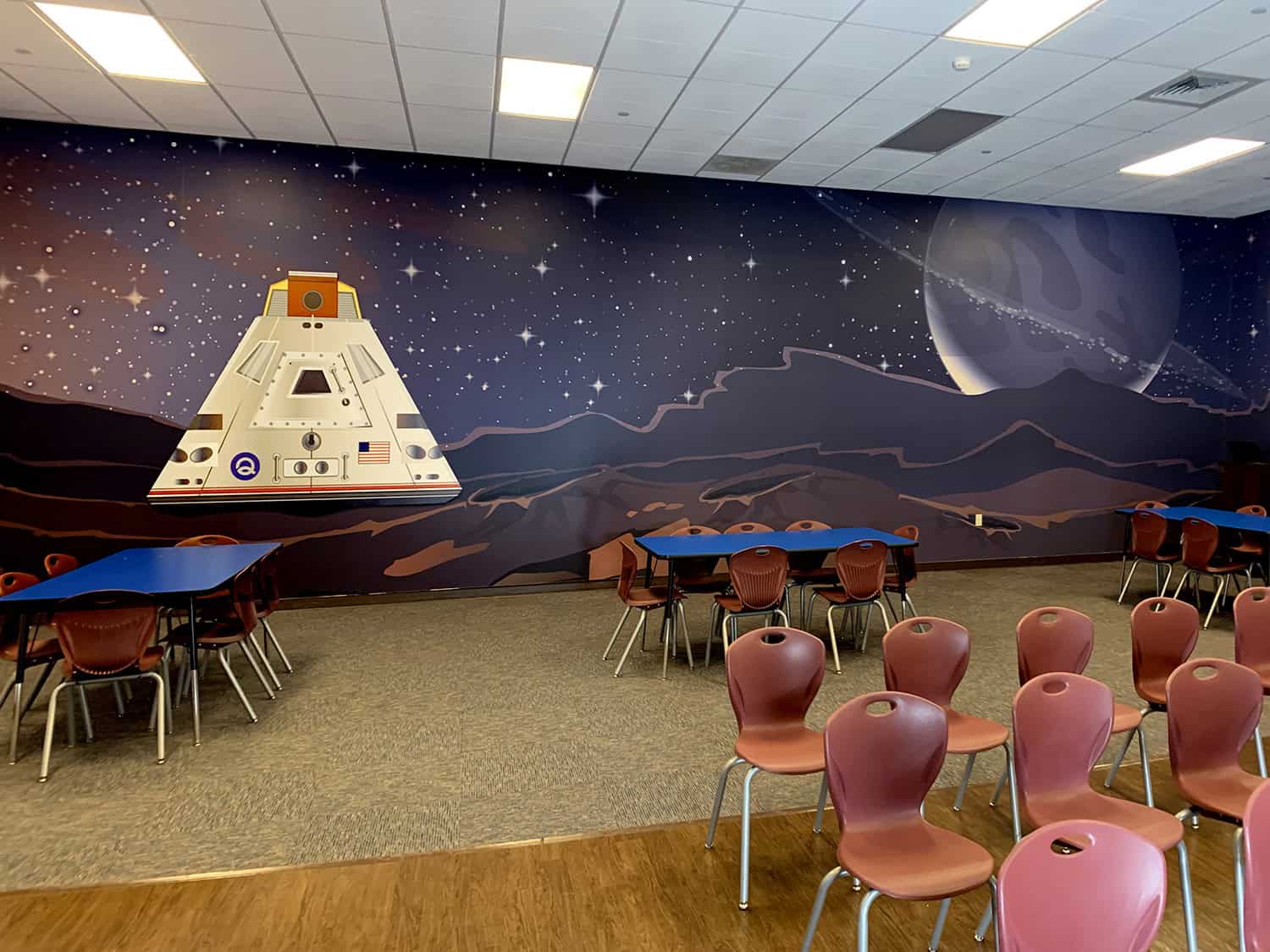 Rocket spaceship mural on space-themed wall in indoor event space, with chairs and tables.