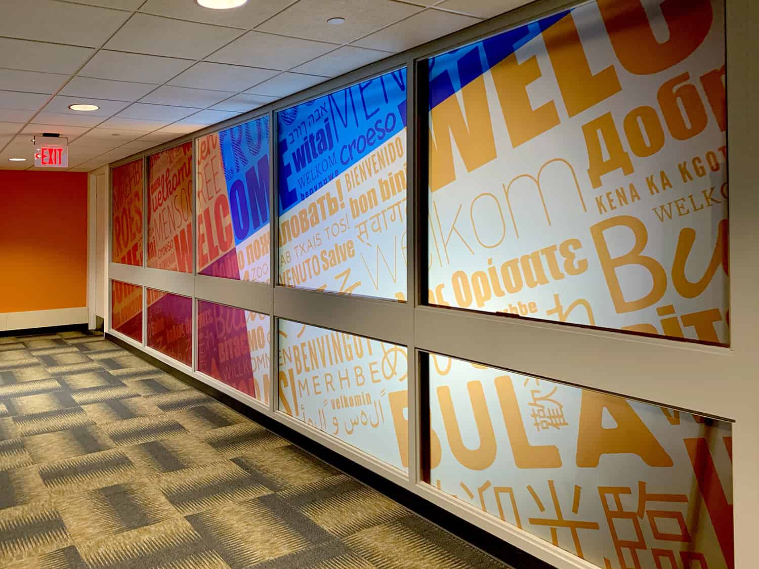 Multilingual welcome sign with vibrant text collage on glass wall in office corridor.