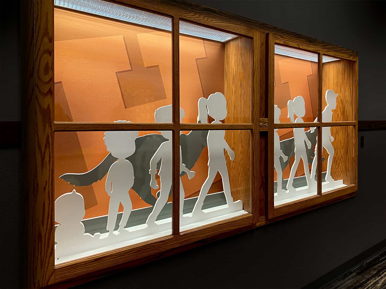 Children and adult silhouettes illuminated inside a wooden display case with backlit paper cutouts.