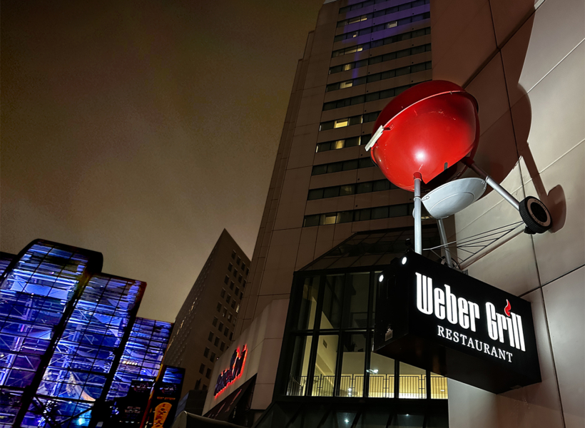 Vibrant red barbecue grill on city sidewalk at night with illuminated Weber Grill Restaurant sign.