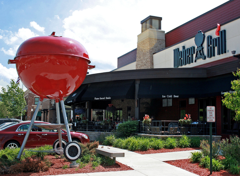 Large red barbecue grill sculpture outside Weber Grill restaurant in sunny weather.