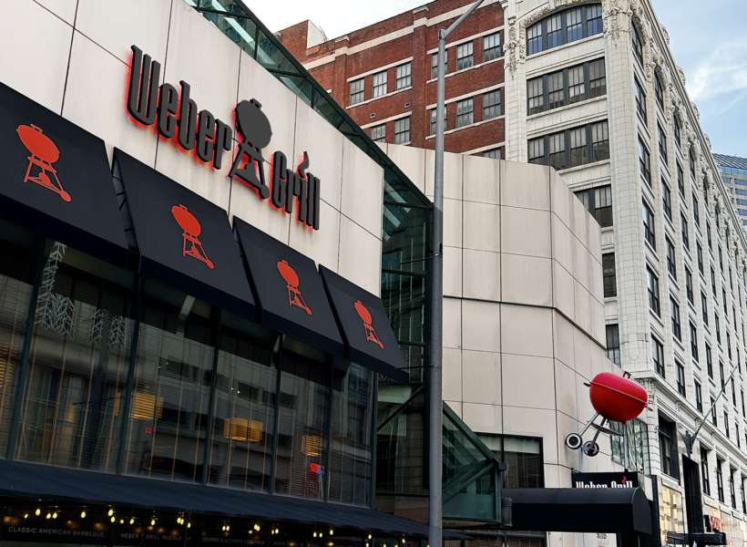 Bright modern outdoor restaurant signage featuring Weber-style barbecue logo, cityscape background.