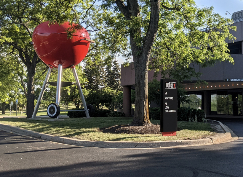 Red BBQ Grill Sculpture outside Weber building in sunny park setting.
